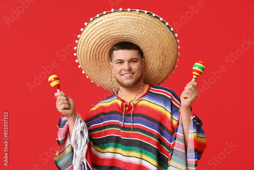 Handsome young happy man in sombrero with maracas on red background. Cinco de Mayo celebration