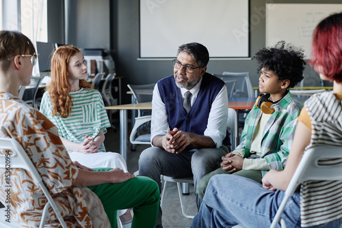 Mature biracial school psychologist talking to group of ethnically diverse teenagers during therapy session