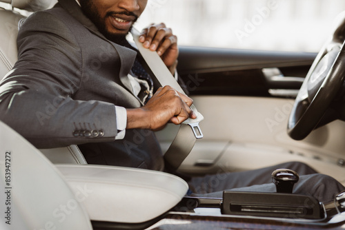 A close-up image of a businessman fastening his seatbelt in the drivers seat of a car. The man is wearing a gray suit and is smiling slightly as he buckles his seatbelt