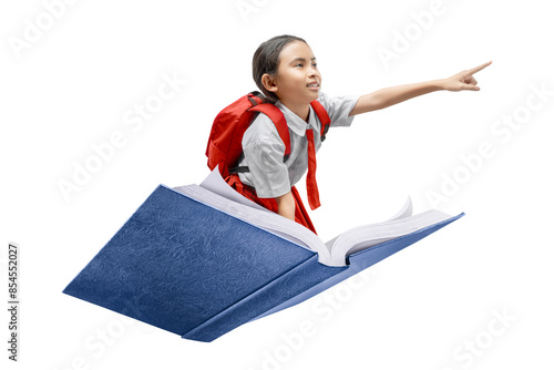 Asian schoolgirl with backpack flying on a book pointing at something with finger on white background