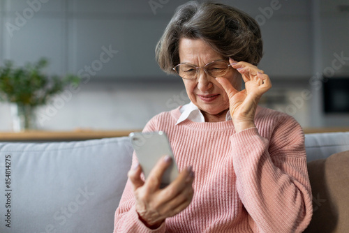 Confused senior woman in eyeglasses squinting eyes, having problems with vision, reading message on smartphone, sitting on couch