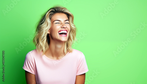 Belle femme aux cheveux blonds, âgée de 25 ans, heureuse et riant aux éclats sur un fond vert, portant un t-shirt rose, image avec espace pour texte.