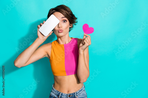 Photo of flirty impressed lady dressed colorful top rising heart modern gadget cover eye empty space isolated turquoise color background