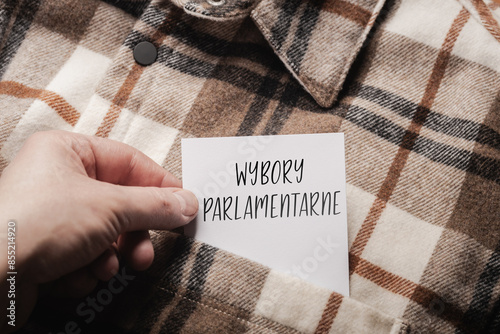 White card with a handwritten inscription "Wybory Parlamentarne", held in the hand against the background of a brown plaid shirt (selective focus), translation: Parliamentary Elections 