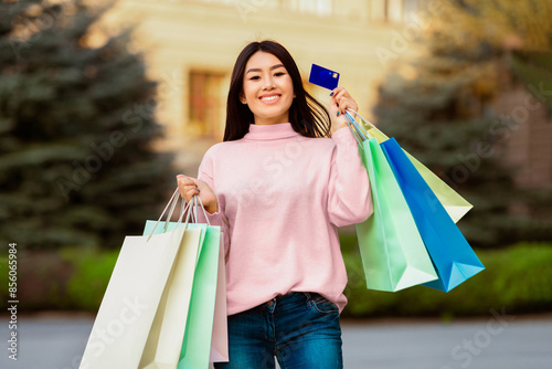 Asian woman is standing holding multiple shopping bags in one hand and a credit card in the other. She appears to be in a shopping area, possibly a mall or retail store, engaged in retail therapy.