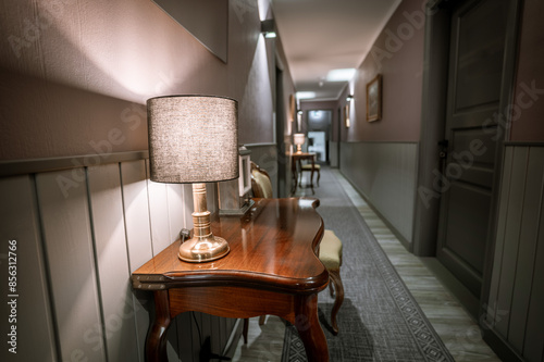 A stylish hotel corridor with elegant wooden tables, lamps, chairs, and patterned carpeting, featuring soft ambient lighting.