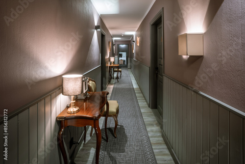 A well-lit, stylish hotel corridor with wooden tables, lamps, chairs, and patterned carpeting, featuring soft wall lighting.