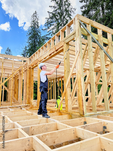 Laborer constructing wooden frame house near forest. Man treating woods, applying fire retardant using sprayer, while dressed in protective suit, helmet. Concept of modern eco-friendly construction.