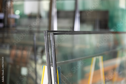 Close-up of insulated glass units stacked on support stand in workshop, with various glass panels and industrial equipment in blurred background