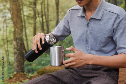 Close up portrait of excited Asian man pouring water from the vacuum flask to the stainless steel mug. Drinking mineral water while trekking in nature forest