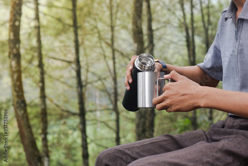 Close up portrait of excited Asian man pouring water from the vacuum flask to the stainless steel mug. Drinking mineral water while trekking in nature forest