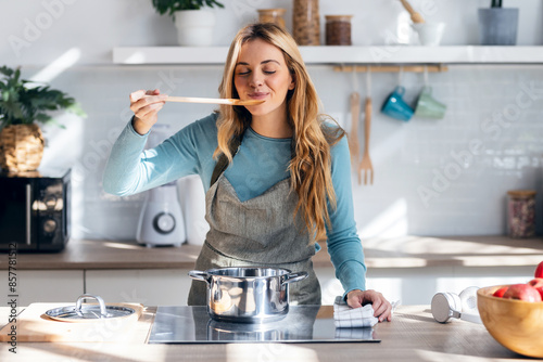 Beautiful woman cooking healthy food in casserole while blowing the spoon to taste the food in the kitchen at home.