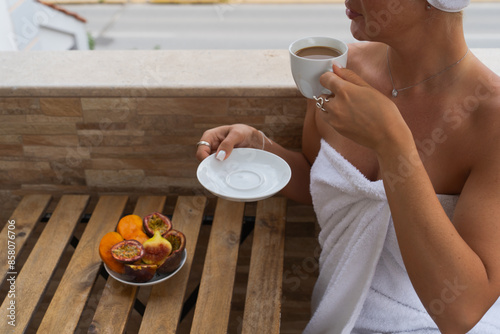 A girl in a white robe drinks coffee on the balcony after a shower, there are fresh cut fruits on the table, summer morning.