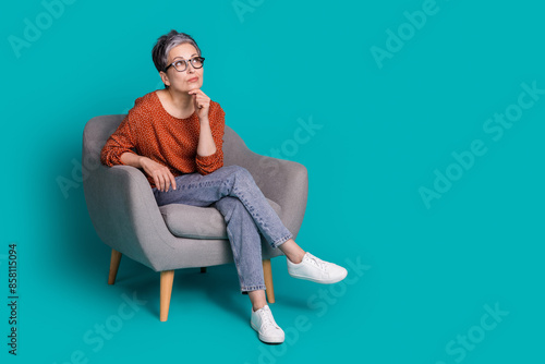 Full body photo of attractive senior woman sit armchair look minded empty space dressed brown clothes isolated on cyan color background