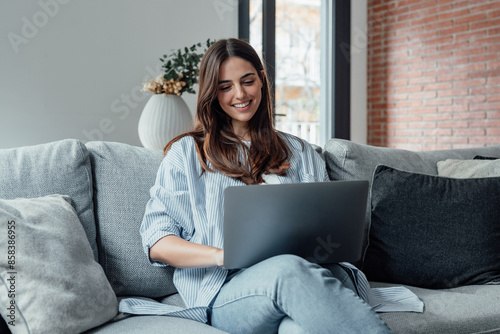 Concentrated millennial girl sit on couch working on laptop browsing internet at home during lazy weekend, focused young woman freelancer busy using computer surfing wireless web shopping online.