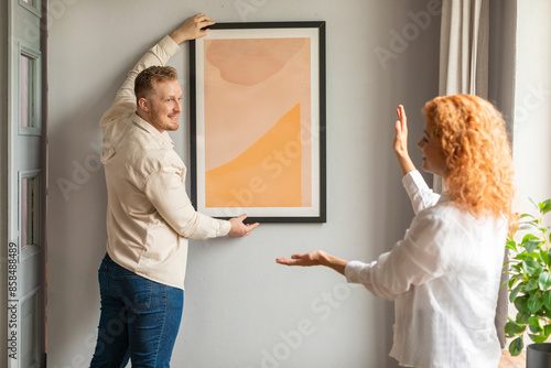 Young spouses arranging new flat, hanging up painting on wall. New home and relocation concept