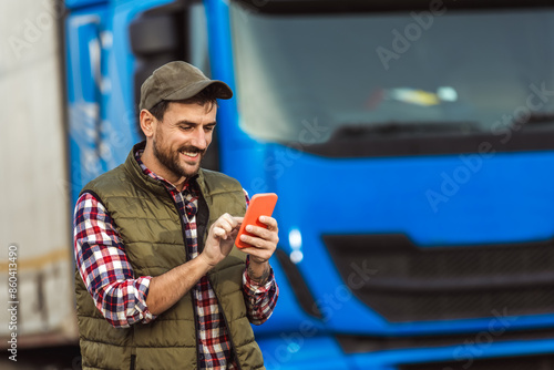 Happy confident male driver standing in front on his truck, using his phone.