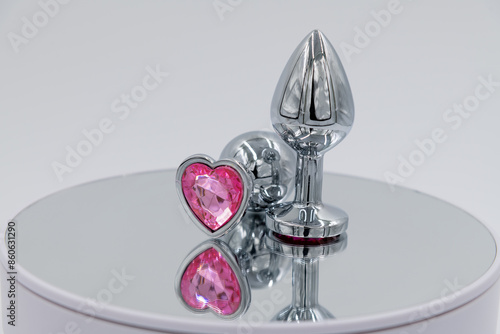 Close-up side view of two metallic butt plugs with pink heart shaped crystal lying on mirror surface against gray background. Soft focus. Adult sex toys theme.