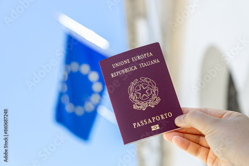 Hand holding an Italian passport with European Union flag waving in the blue sky in the background