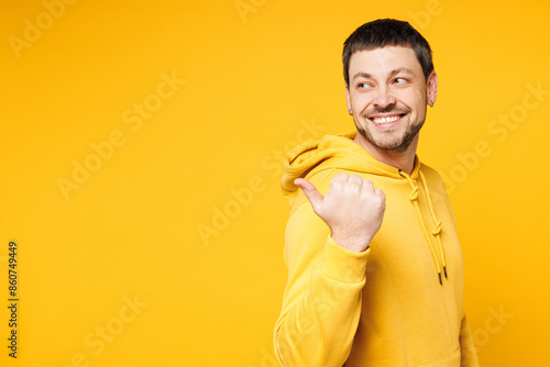 Side view young smiling happy man wear hoody casual clothes point thumb finger aside on area mockup workspace look camera isolated on plain yellow orange background studio portrait. Lifestyle concept