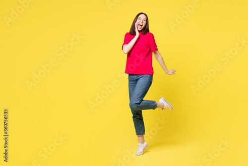 Full body young smiling happy charming nice woman wear pink t-shirt casual clothes stand put hand on face raise up leg isolated on plain yellow orange background studio portrait. Lifestyle concept.
