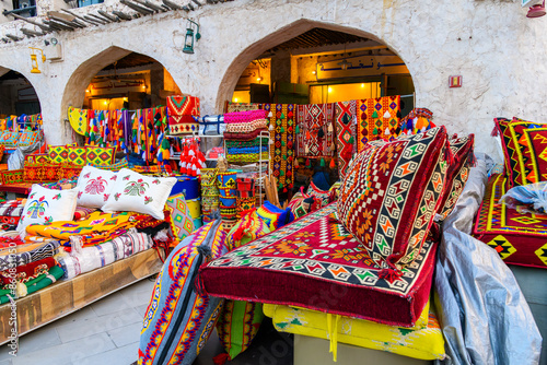 A colorful outdoor storefront display selling vibrantly colored fabrics on blankets, rugs and pillows in the Souq Waqif marketplace of Doha, Qatar. 