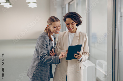 Stylish busy female coworkers discussing project standing in modern office and use digital tablet