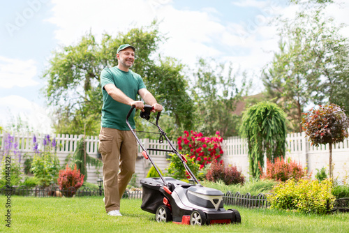 Positive man mowing the lawn using an electric lawn mower on backyard near country house, full length, free space