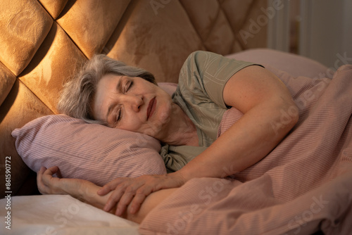 At night, an elderly woman can be seen sleeping soundly in bed. She is covered with a blanket, and her arms are wrapped around a pillow. The bed is made with pink sheets and a pink blanket