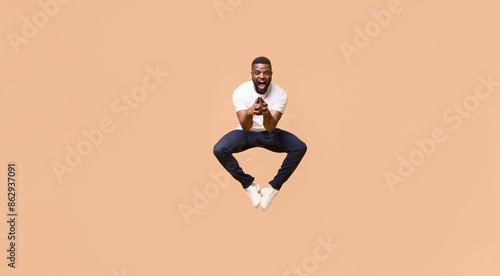 A young Black man jumps in mid-air with a wide smile and open mouth, his arms bent at the elbows, as if holding something. He is wearing a white t-shirt and blue jeans