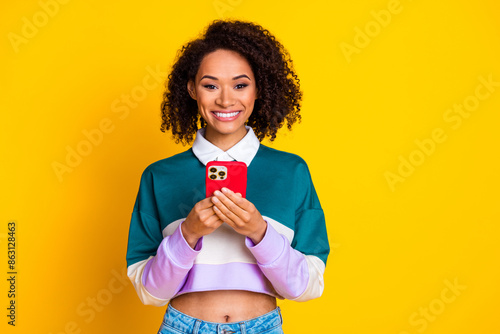 Photo of cheerful woman with curly hairstyle dressed striped top holding smartphone in hands smiling isolated on yellow color background