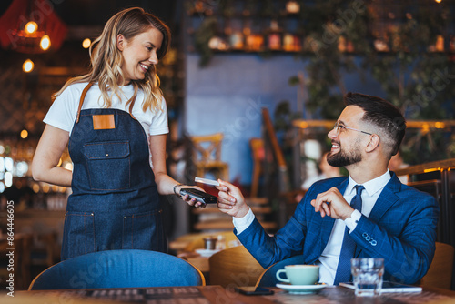 Smiling man in a suit engages with a waitress, using a card for payment, reflecting a casual yet professional dining transaction in a cafe setting.