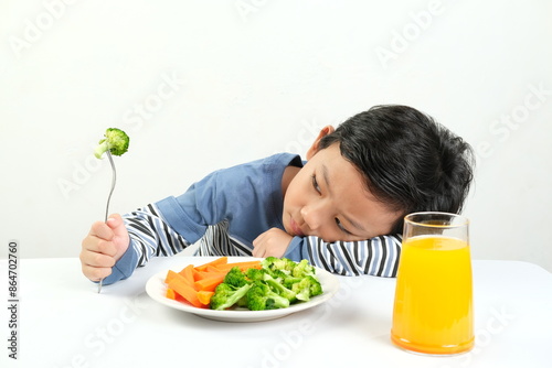 A young boy leans on the table, refusing to eat his vegetables and drink orange juice, displaying boredom and reluctance. Perfect for illustrating picky eating habits in children.