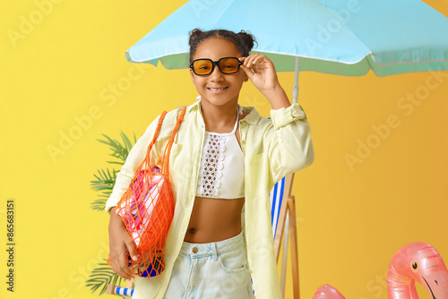 Happy African-American girl with beach accessories and umbrella on yellow background