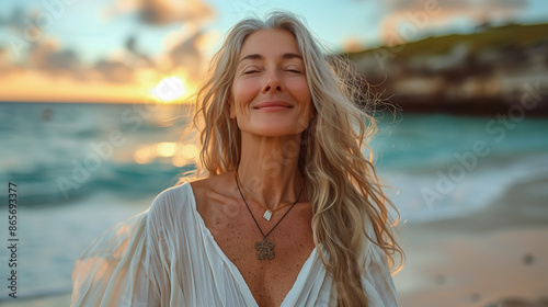 A woman with long, silver hair stands on a beach, her eyes closed in peaceful contentment as the sun sets behind her