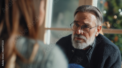 A mature man with glasses and a beard speaks to a young woman in a living room. They are having a conversation.