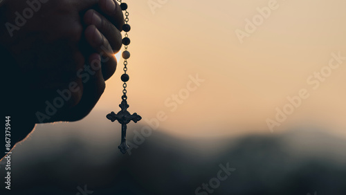 Man's hands pray to hold a bead rosary with Jesus Christ on the cross or the Crucifix with sunset. Christian Catholic religious faith holding a black rosary and praying.