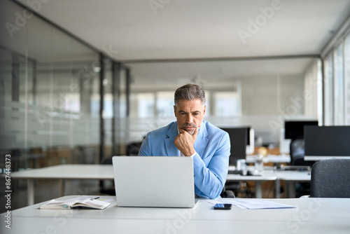 Busy serious middle aged professional business man financial investor, 50 year old senior businessman executive wearing suit using laptop computer working on investment project sitting at office desk.