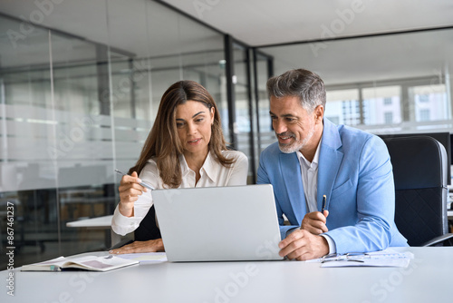 Two busy happy middle aged professional business man and woman executive leaders team using laptop working on computer at work desk having conversation on financial project at meeting in office.