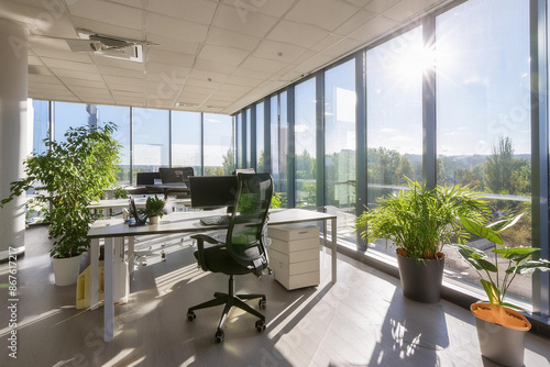 A stylish, minimalist office space with large windows, sleek furniture, and vibrant indoor plants. The scene is brightly lit by natural sunlight, creating an inviting workspace