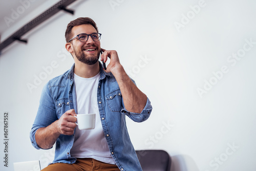 Handsome bearded man leaning on office desk, drinking coffee and talking on the phone.