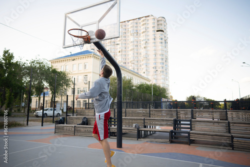 man in a grey hoodie and red shorts prepares to execute a basketball layup on an outdoor court. The basketball is about to enter the hoop