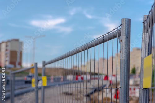 Ogrodzenie z siatki metalowej terenu na budowie. Błękitne niebo nad placem budowy dróg w mieście. W tle widoczne bloki mieszkalne, w pobliżu ogrodzenie z siatki stalowej.