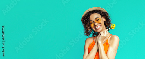 Black woman with curly brown hair smiles at the camera while holding a yellow lollipop in her right hand. She is wearing a straw hat, orange tank top, and yellow heart-shaped sunglasses.