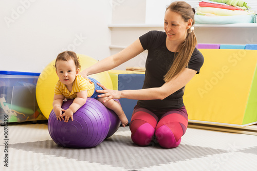 Happy child sitting on a violet fitness ball with his physiotherapist during a session of physical rehabilitation and exercise. Physical rehabilitation of children and early intervention