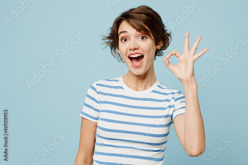 Young happy surprised shocked woman she wears striped t-shirt casual clothes showing okay ok gesture look camera isolated on plain pastel light blue cyan background studio portrait. Lifestyle concept.