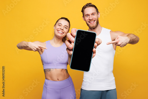 Young strong fitness trainer sporty two man woman wear blue clothes spend time in home gym hold use show blank screen mobile cell phone isolated on plain yellow background. Workout sport fit concept.
