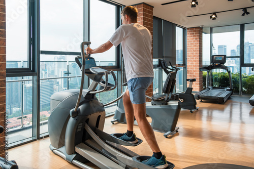 Man working out on an elliptical trainer in modern gym with city view. Male doing cardio training on elliptical machine in fitness studio in residental building. Fitness, wellness, healthy lifestyle