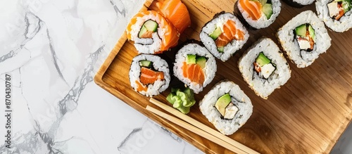 A wooden plate displays a variety of tasty sushi rolls, with chopsticks, against a plain background in the copy space image.