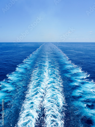 Wide turbulent white foam water trail behind a large cruise ship in a calm blue sea on a sunny day